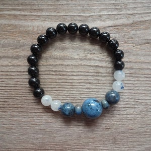 Op de afbeelding: Een zwart-wit geperst armband met een grote blauwe edelsteen in het midden. De armband is gemaakt met zwarte onyx kralen en witte maansteen kralen.