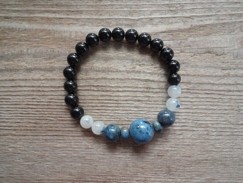 Op de afbeelding: Een zwart-wit geperst armband met een grote blauwe edelsteen in het midden. De armband is gemaakt met zwarte onyx kralen en witte maansteen kralen.