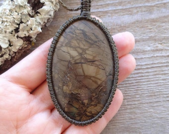 May include: An oval-shaped pendant featuring a brown, cracked-pattern stone encased in a dark olive green macrame cord setting. The pendant is attached to a matching cord necklace, showcasing intricate knot work around the stone's perimeter.