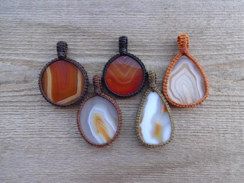 May include: Five agate stone pendants with different patterns and colors, each wrapped in a macrame cord. The pendants are in shades of brown, orange, and white.