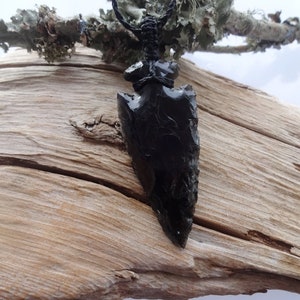 May include: Black obsidian arrowhead pendant on a brown wooden surface. The arrowhead is tied with black cord.