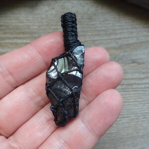 May include: A raw, dark, reflective stone pendant encased in a black macrame cord netting. The stone features a smooth, glassy texture with subtle gray and black striations. A thick, braided black cord loop extends from the top for suspension.