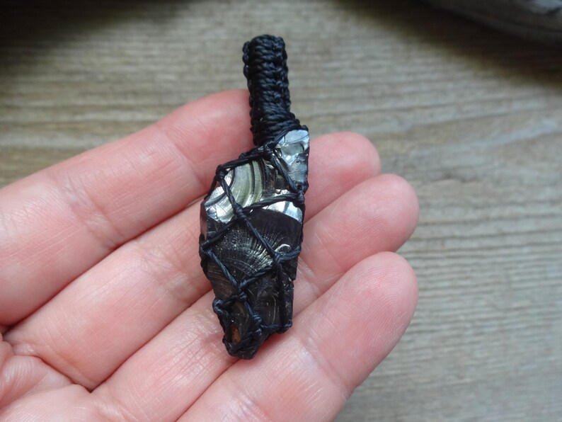 May include: A raw, dark, reflective stone pendant encased in a black macrame cord netting. The stone features a smooth, glassy texture with subtle gray and black striations. A thick, braided black cord loop extends from the top for suspension.