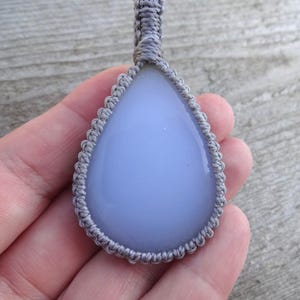 Peut inclure: Pendentif en forme de goutte d'eau avec une pierre bleu p&acirc;le, sertie dans un motif macram&eacute; gris. Le pendentif est suspendu &agrave; un cordon gris assorti. La pierre a une surface lisse et polie.