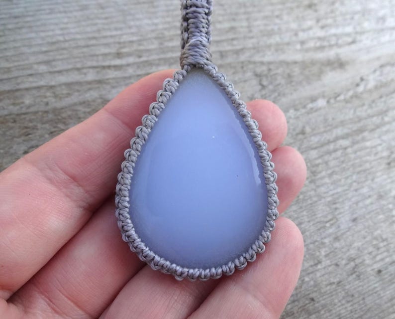 Peut inclure: Pendentif en forme de goutte d'eau avec une pierre bleu p&acirc;le, sertie dans un motif macram&eacute; gris. Le pendentif est suspendu &agrave; un cordon gris assorti. La pierre a une surface lisse et polie.