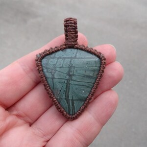 May include: A green and brown stone pendant with a macrame cord design. The pendant is in the shape of a triangle and has a brown cord loop at the top for hanging.