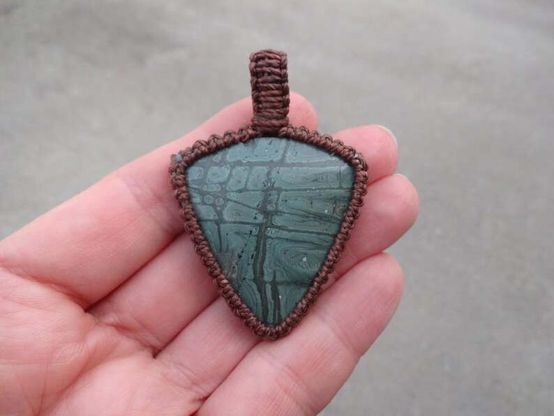 May include: A green and brown stone pendant with a macrame cord design. The pendant is in the shape of a triangle and has a brown cord loop at the top for hanging.