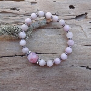 May include: A pink and white gemstone bracelet with a larger pink gemstone in the center. The bracelet is made of round beads with small silver accents.
