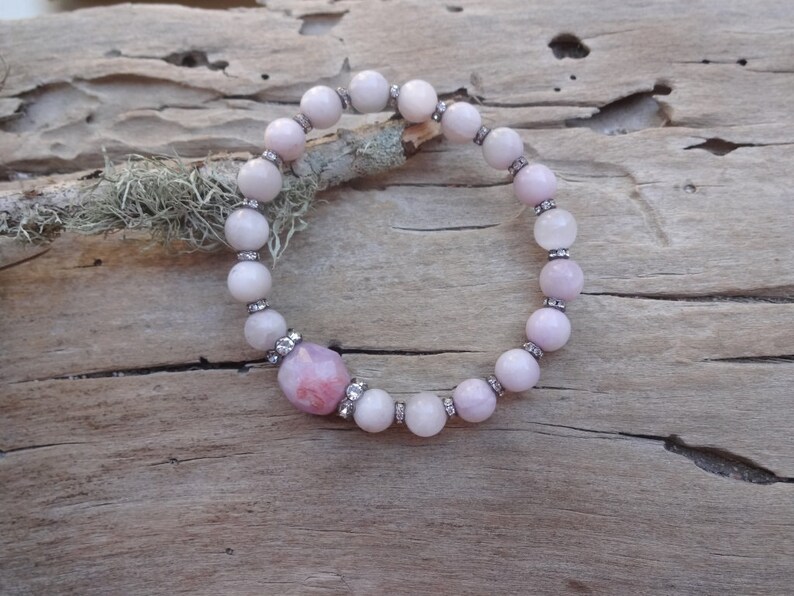 May include: A pink and white gemstone bracelet with a larger pink gemstone in the center. The bracelet is made of round beads with small silver accents.