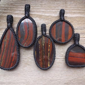 May include: Five pendants with red and brown striped stones, each encased in a black woven macrame setting. The pendants vary in shape, including oval, teardrop, and round designs. The pendants are displayed on a wooden surface.