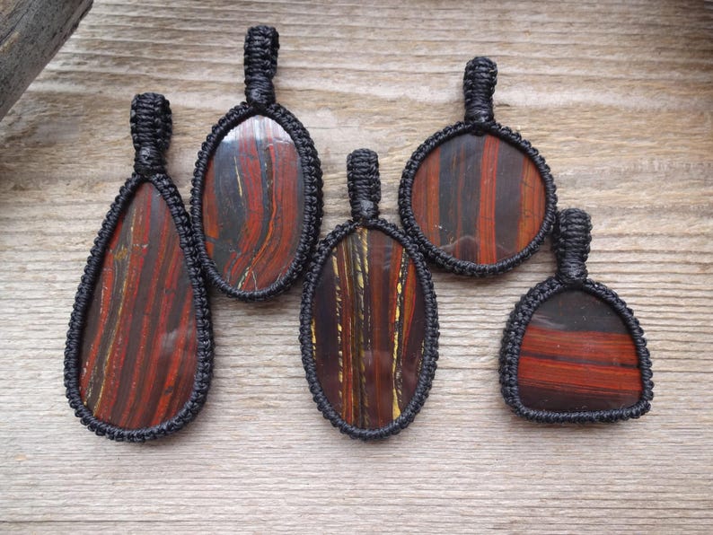May include: Five pendants with red and brown striped stones, each encased in a black woven macrame setting. The pendants vary in shape, including oval, teardrop, and round designs. The pendants are displayed on a wooden surface.