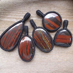 May include: A collection of six tiger eye pendants with black macrame detailing. The pendants vary in shape, showcasing the natural red and brown striped patterns of the stone. They are displayed on a wooden surface.