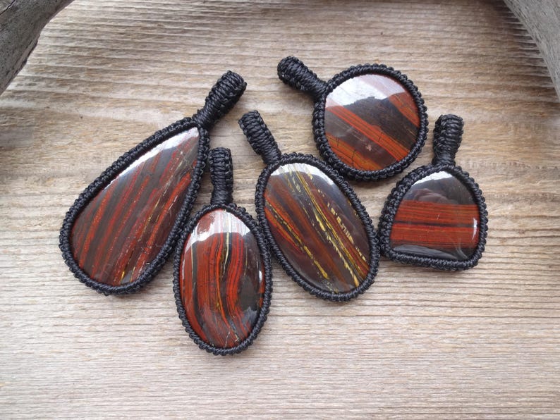 May include: A collection of six tiger eye pendants with black macrame detailing. The pendants vary in shape, showcasing the natural red and brown striped patterns of the stone. They are displayed on a wooden surface.