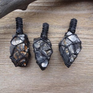 May include: Three black crystal pendants with a woven black cord. The crystals are encased in a black net-like material. The background is a light brown wooden surface with a piece of driftwood.