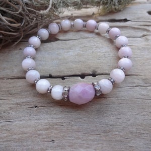 May include: A pink and white beaded bracelet with a larger pink gemstone in the center. The bracelet is made of round beads with a silver accent on each bead.