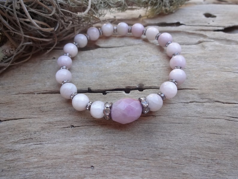 May include: A pink and white beaded bracelet with a larger pink gemstone in the center. The bracelet is made of round beads with a silver accent on each bead.