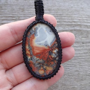 May include: A close-up of an oval-shaped pendant with a black cord. The pendant is made of a red, white, and gray stone with a swirling pattern.