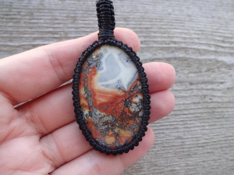 May include: A close-up of an oval-shaped pendant with a black cord. The pendant is made of a red, white, and gray stone with a swirling pattern.