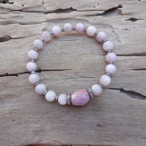 May include: A pink and white beaded bracelet with a larger pink gemstone in the center. The bracelet is on a wooden surface.