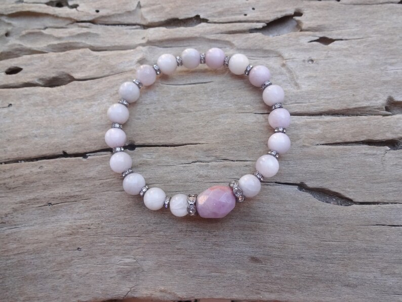 May include: A pink and white beaded bracelet with a larger pink gemstone in the center. The bracelet is on a wooden surface.