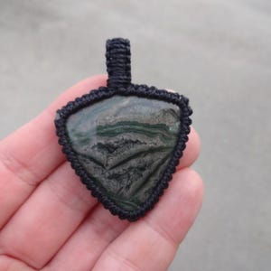 May include: A black cord pendant with a triangular green and black stone. The stone has a swirling pattern.