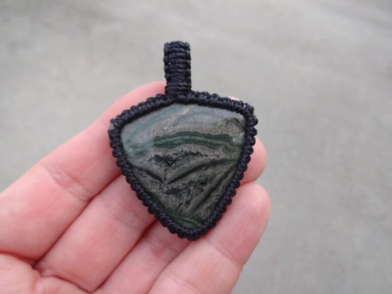 May include: A black cord pendant with a triangular green and black stone. The stone has a swirling pattern.