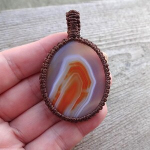 May include: A brown cord wrapped around an oval agate pendant with white and orange banding.