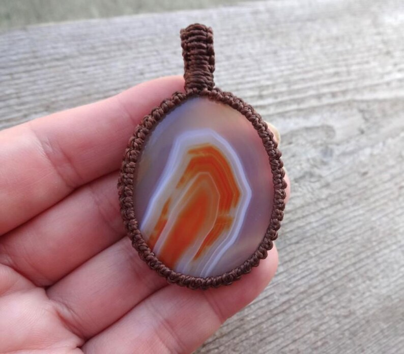 May include: A brown cord wrapped around an oval agate pendant with white and orange banding.