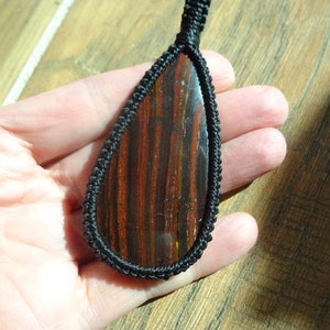 May include: A teardrop-shaped pendant featuring a polished stone with red and brown stripes, encased in a black woven cord. The pendant is suspended from a matching black cord, showcasing a natural gemstone.