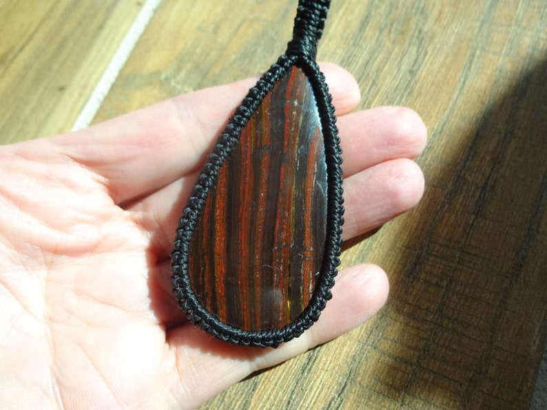 May include: A teardrop-shaped pendant featuring a polished stone with red and brown stripes, encased in a black woven cord. The pendant is suspended from a matching black cord, showcasing a natural gemstone.