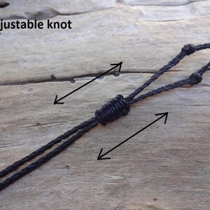 May include: A black cord featuring an adjustable knot mechanism. The cord is composed of two strands that pass through a central sliding knot, allowing for length modification. The text "adjustable knot" is visible in the upper left corner.