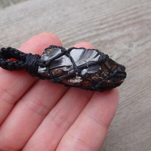 May include: A dark, metallic-looking stone pendant wrapped in black macrame. The stone has a shiny, reflective surface with a rough, uneven texture. The macrame cord forms a net around the stone, with a loop for hanging.