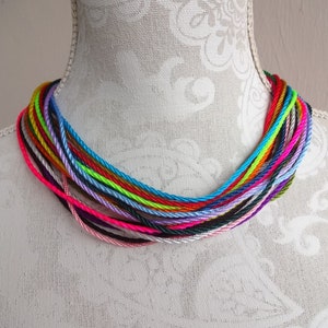 May include: A colorful necklace made of multiple strands of thin, braided cord in various colors, including pink, purple, blue, green, yellow, orange, and red. The necklace is displayed on a white mannequin.