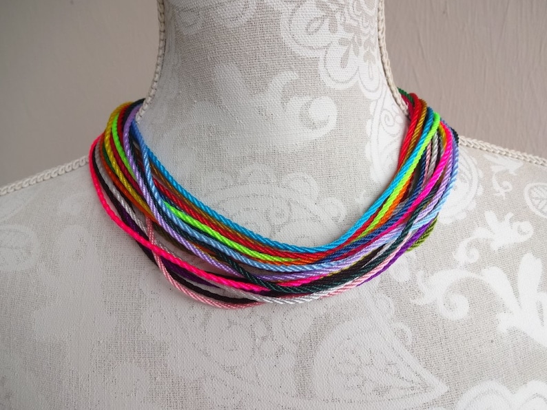 May include: A colorful necklace made of multiple strands of thin, braided cord in various colors, including pink, purple, blue, green, yellow, orange, and red. The necklace is displayed on a white mannequin.