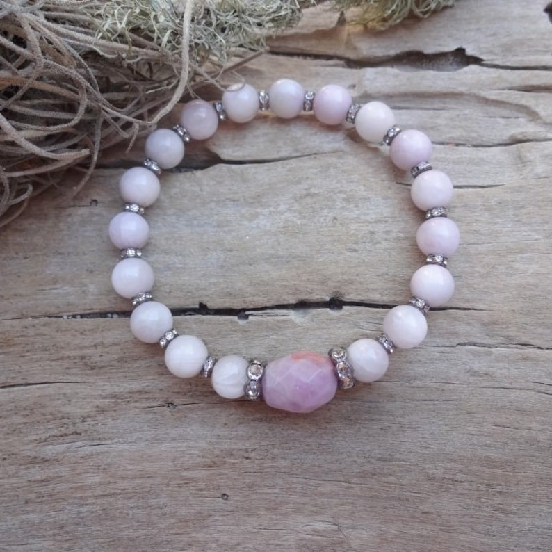 May include: A pink and white gemstone bracelet with a larger pink gemstone in the center. The bracelet is made of small round beads with silver accents.