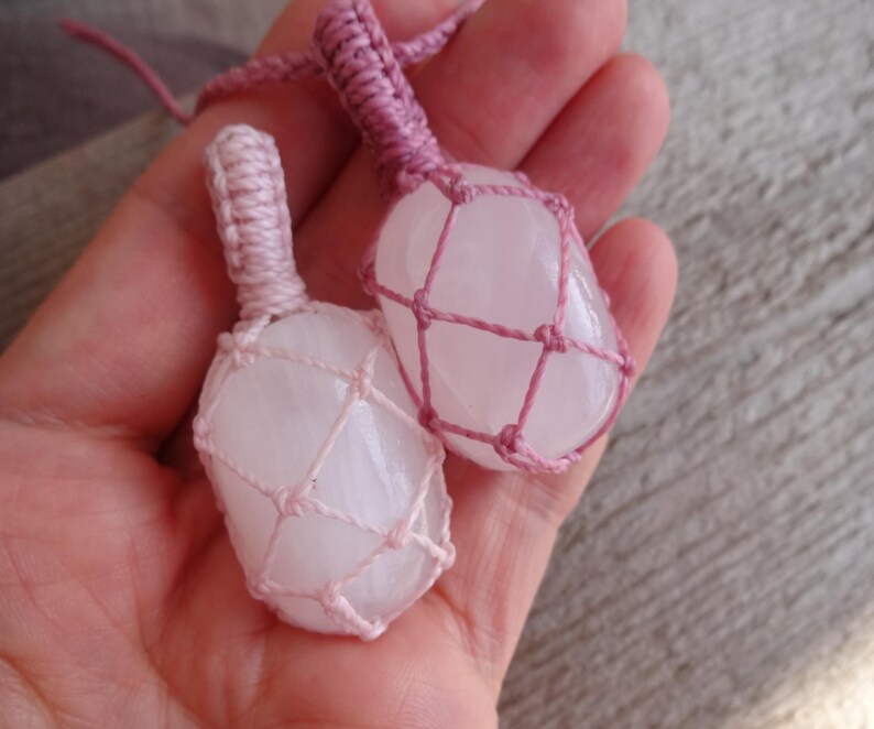 May include: Two pink crystal stones are wrapped in pink macrame cord. The stones are smooth and have a natural, cloudy appearance.