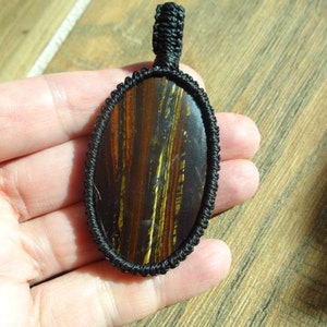 May include: Oval pendant with a polished tiger's eye stone, featuring brown, gold, and red striations. The stone is encased in a black woven macrame setting with a loop for hanging. The pendant is held in a hand.