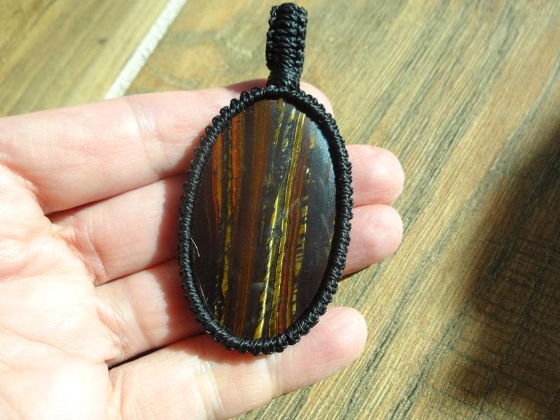 May include: Oval pendant with a polished tiger's eye stone, featuring brown, gold, and red striations. The stone is encased in a black woven macrame setting with a loop for hanging. The pendant is held in a hand.