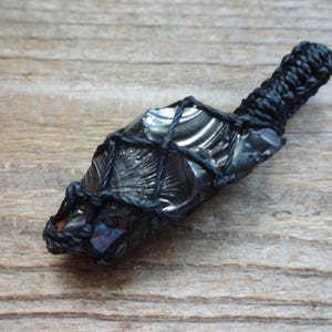 May include: A dark, reflective obsidian stone pendant encased in a black macrame cord netting. The stone features a smooth, glossy surface with subtle iridescent flashes of blue and purple, secured by a braided black cord loop at the top.