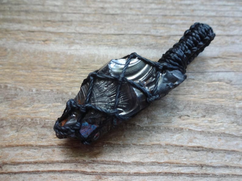May include: A dark, reflective obsidian stone pendant encased in a black macrame cord netting. The stone features a smooth, glossy surface with subtle iridescent flashes of blue and purple, secured by a braided black cord loop at the top.