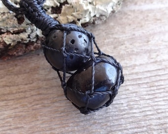 May include: Two black lava rock beads are encased in a black macrame net. The beads are tied together with a black cord, which is knotted at the top to form a loop for hanging.