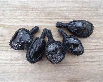 May include: Five black obsidian pendants with a woven black cord. The obsidian stones have a polished, reflective surface and are set in a macrame-style weave. The pendants are displayed on a light brown wooden surface.