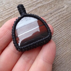May include: A dark, triangular pendant with a polished, reflective surface, encased in a black woven macrame setting. The pendant has a loop at the top for hanging. The background is a wooden surface.