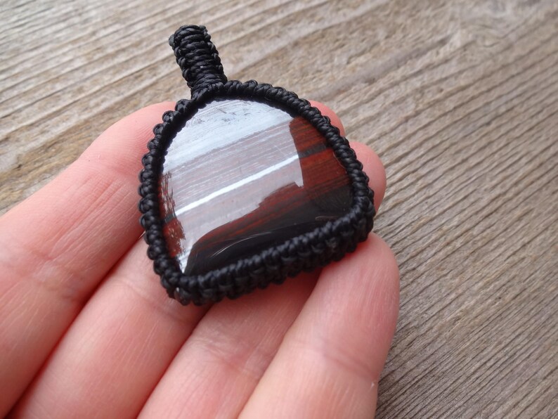 May include: A dark, triangular pendant with a polished, reflective surface, encased in a black woven macrame setting. The pendant has a loop at the top for hanging. The background is a wooden surface.