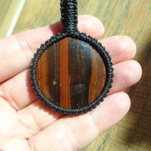 May include: A round pendant with a polished stone center, featuring vertical bands of reddish-brown and dark brown. The stone is encased in a black macrame setting with a matching black cord for wearing. The pendant is held in a hand.