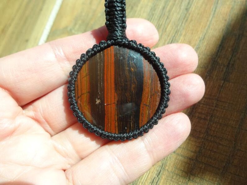 May include: A round pendant with a polished stone center, featuring vertical bands of reddish-brown and dark brown. The stone is encased in a black macrame setting with a matching black cord for wearing. The pendant is held in a hand.