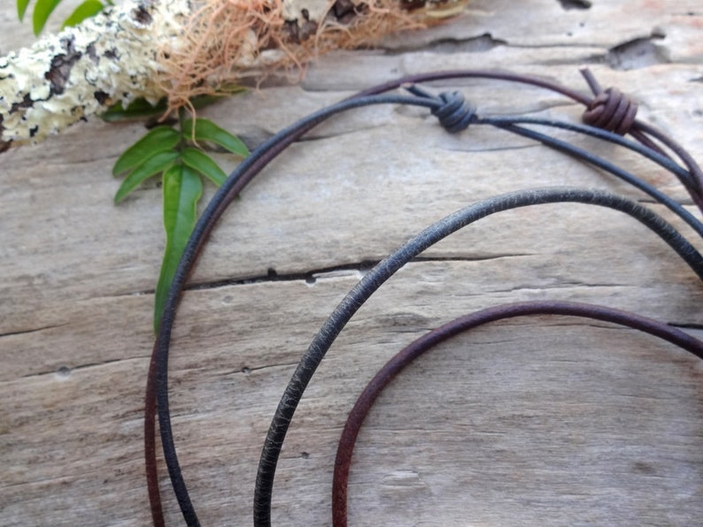 May include: Two leather cords, one black and one brown, are tied together with a knot. The cords are lying on a wooden surface.
