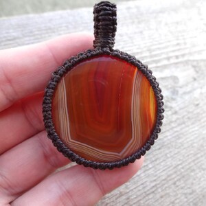 May include: A brown cord wrapped around a round, red, white, and orange agate pendant. The pendant is hanging from a brown cord.