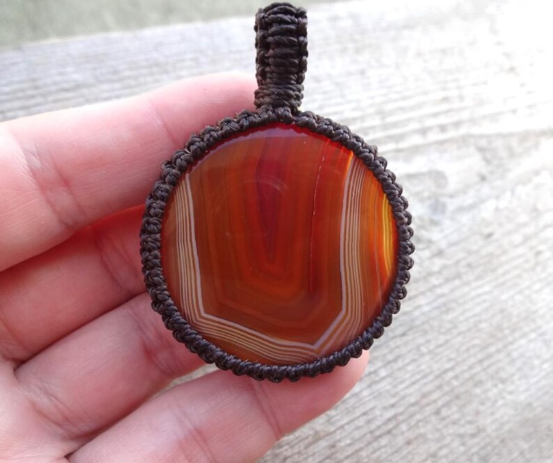 May include: A brown cord wrapped around a round, red, white, and orange agate pendant. The pendant is hanging from a brown cord.