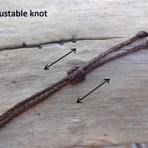 May include: Close-up of a brown cord with an adjustable knot. The text "adjustable knot" is visible at the top of the image. The cord is made of a woven material and is set against a wooden background.